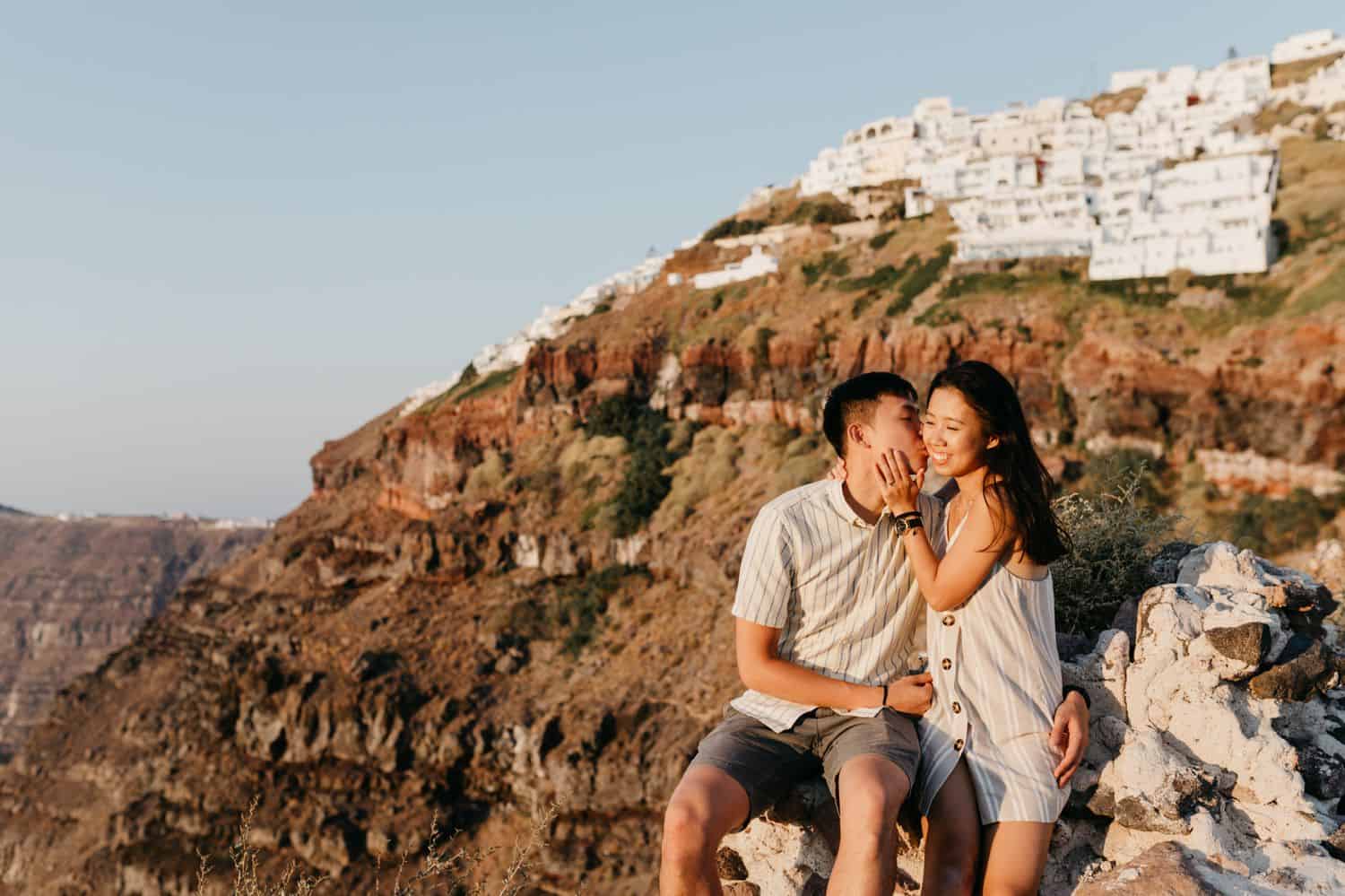 A couple sits on the side of a cliff on a sunny day in Greece