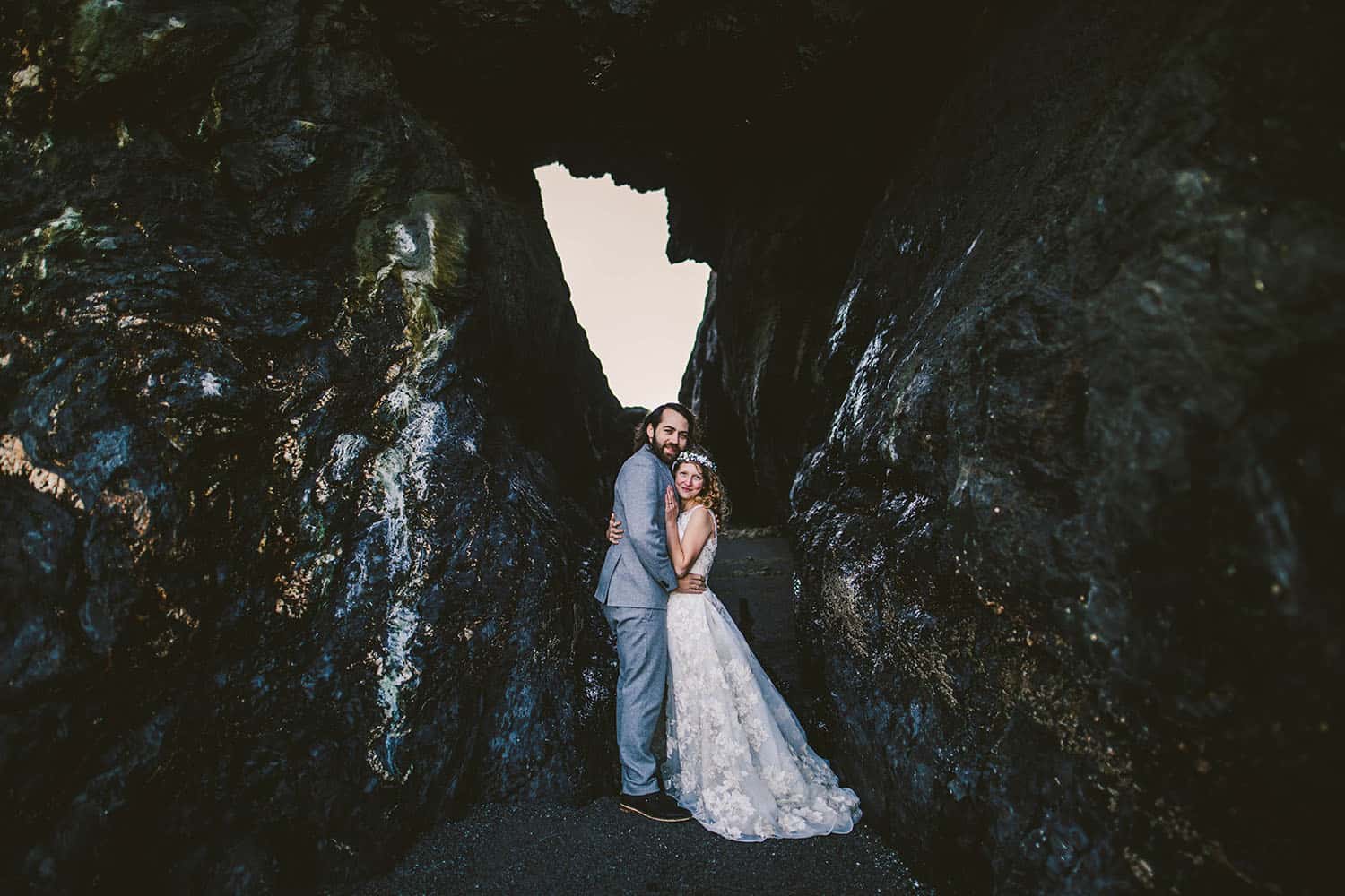 Color full-length portrait of a caucasion bride and groom embracing at the entrace to a sea cave. The groom has a brown beard and is wearing a gray suit, and the bride is wearing a flower crown over her blonde curls.