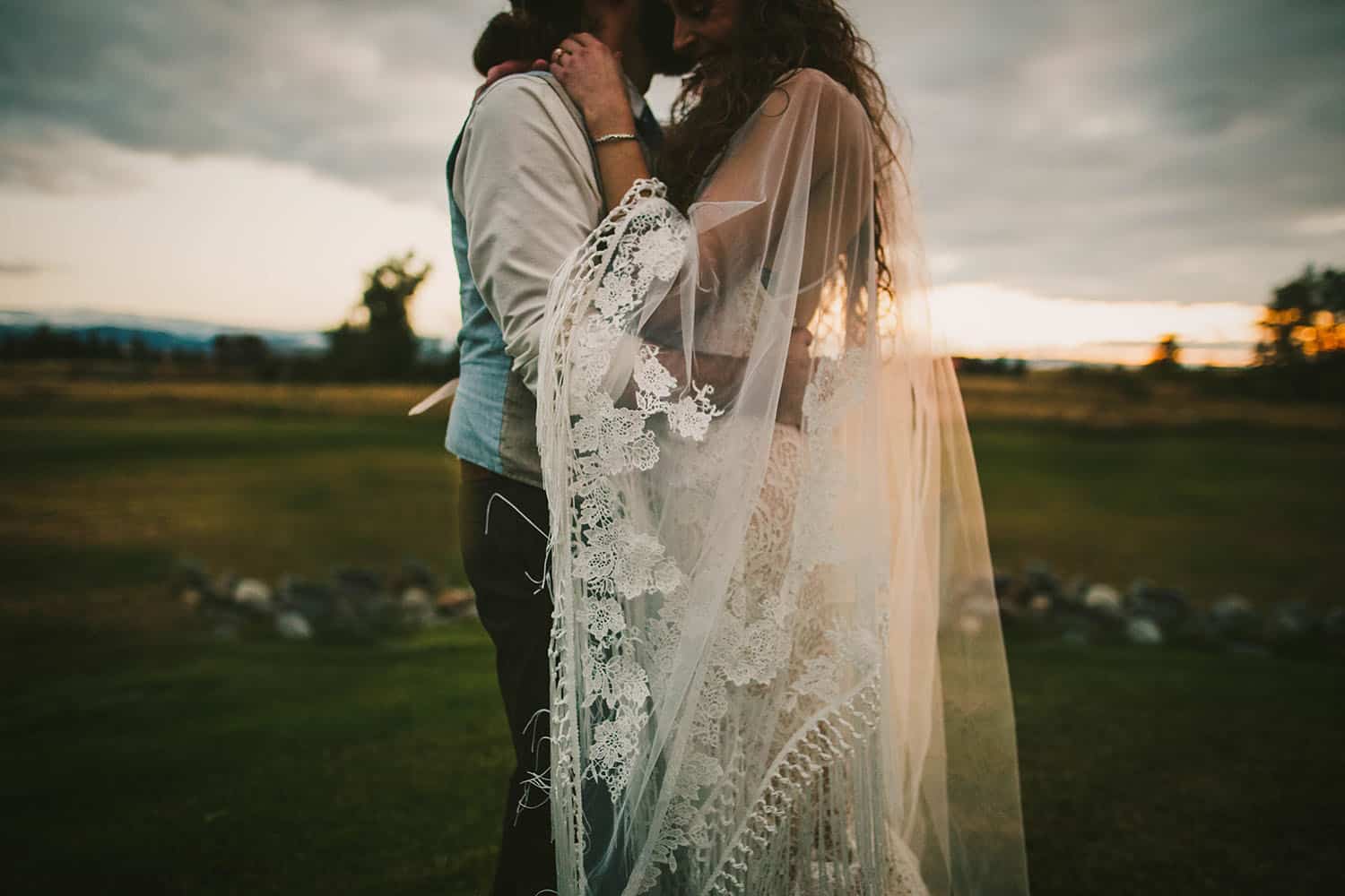 Color detail image of a caucasian bride and groom embracing in a field at dusk. The couple are only visible from their nose to their knees, with the primary focus on the bride's handmade lace veil that wraps around them both.