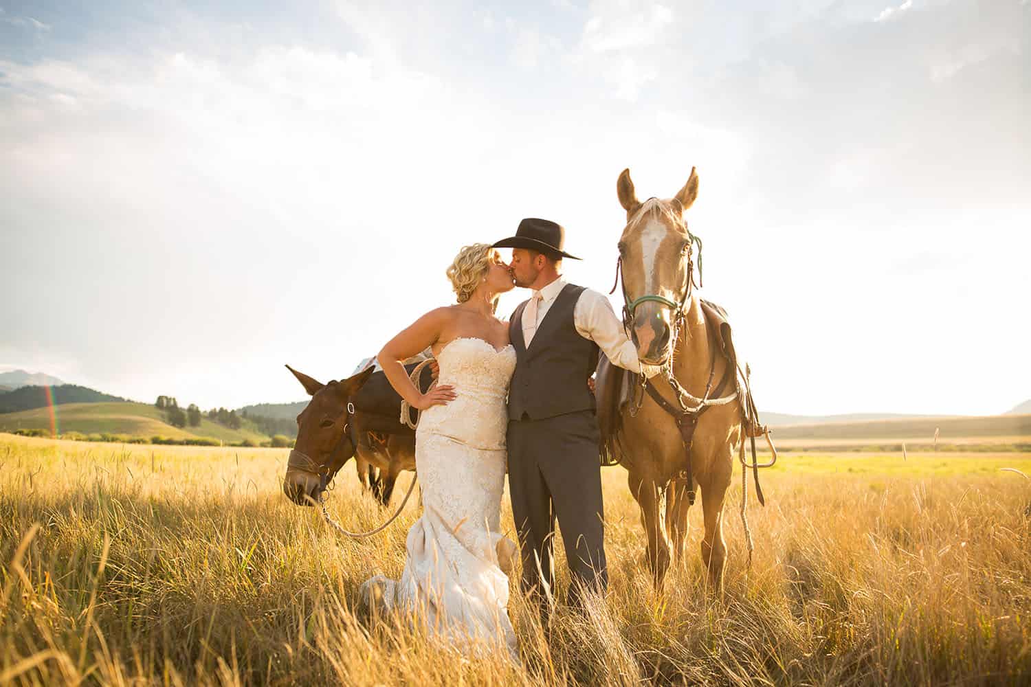 Color photo of a blonde bride kissing her cowboy-hatted groom in a field at midday. The two stand with their horses.