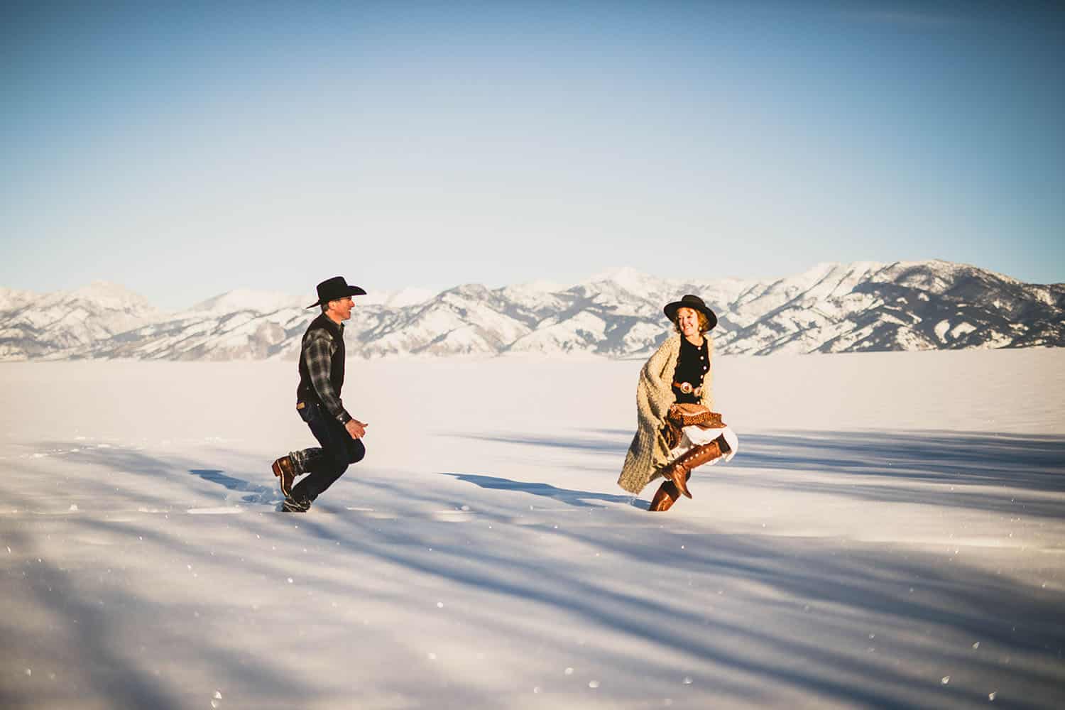 Color image of a caucasion couple in boots and cowboy hats tromping through a snowy landscape in the late afternoon.