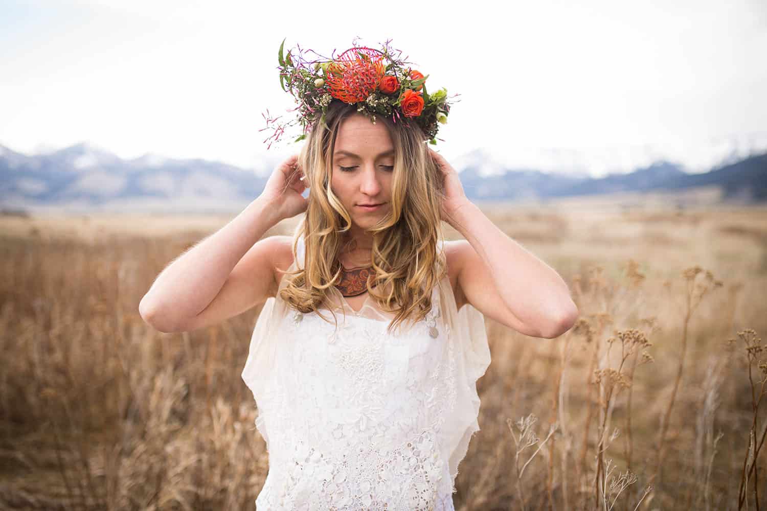 Color half-length photograph of a blonde bride wearing white overalls, a veil, and a flower crown, standing in open shade in a large overgrown field with mountains in the background.