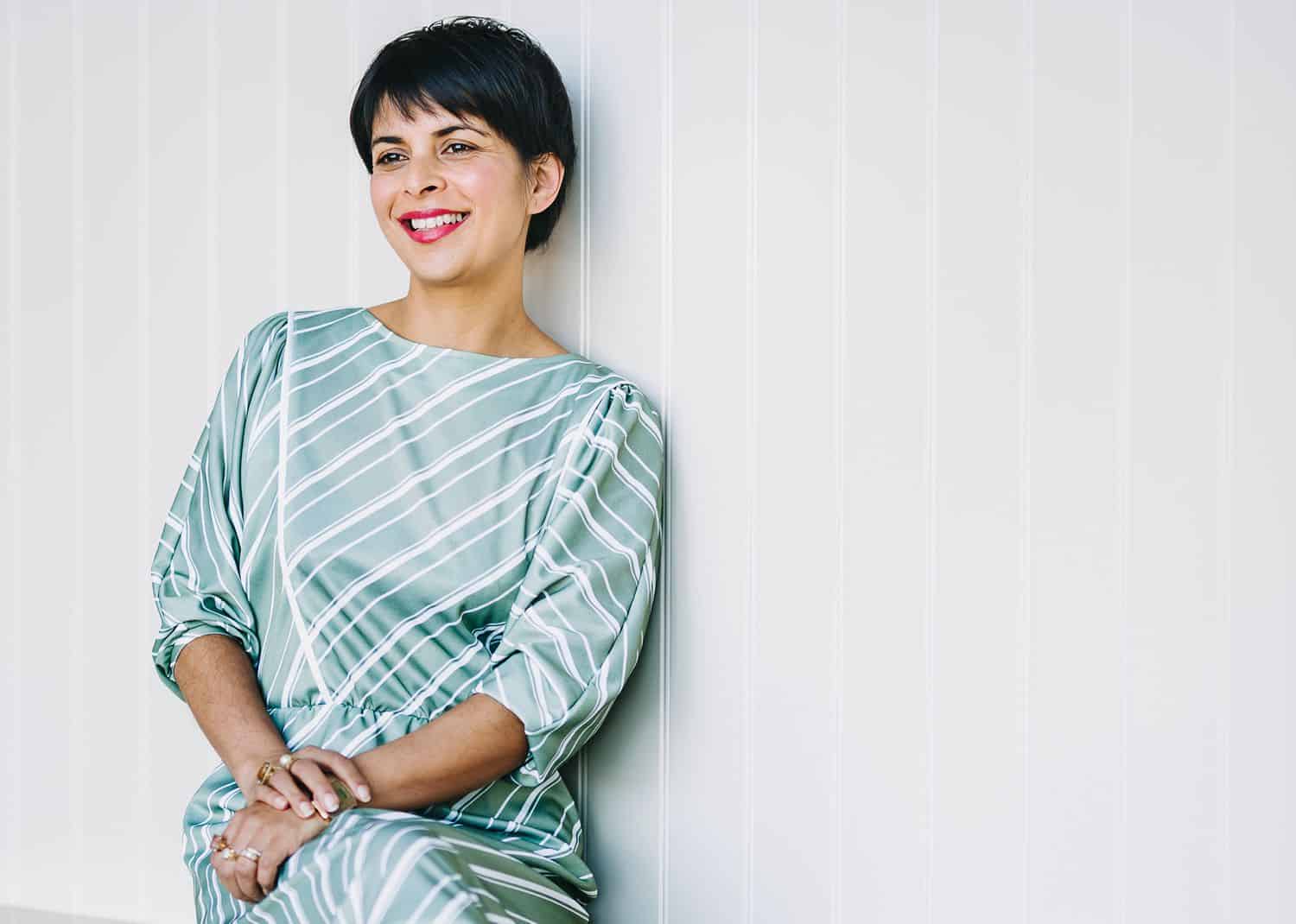 A brunette woman with a pixie haircut leans against a white paneled wall wearing a blue silk dress