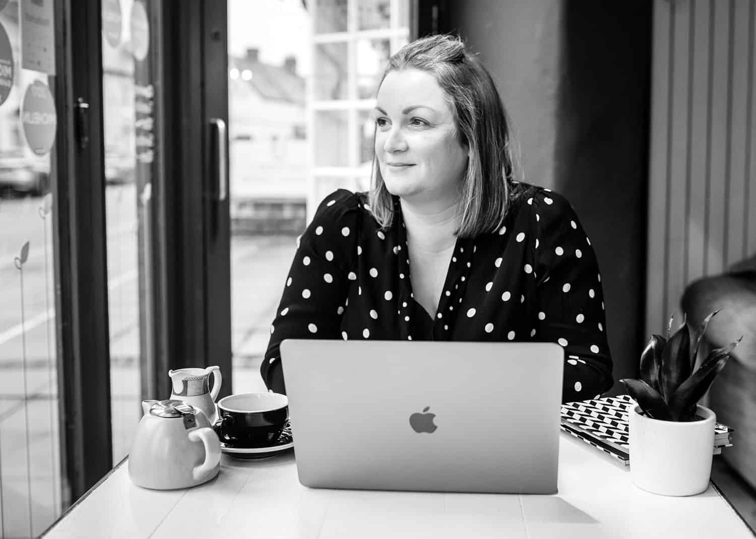 A blonde woman in a black polka-dot dress sits at her laptop in a coffee shop drinking tea