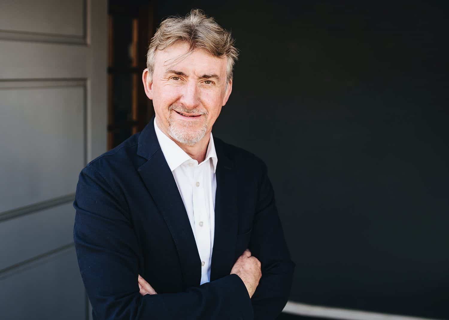 A gentleman in a white shirt and black suit jacket stands in front of an open doorway with his arms folded