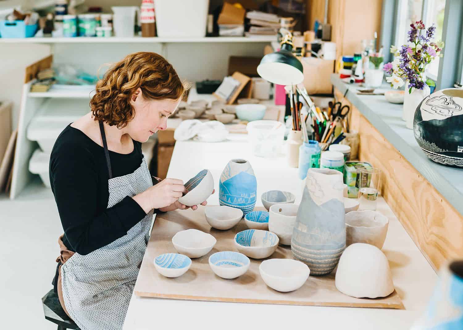 A woman with red hair sits in an apron at a pottery table painting vases