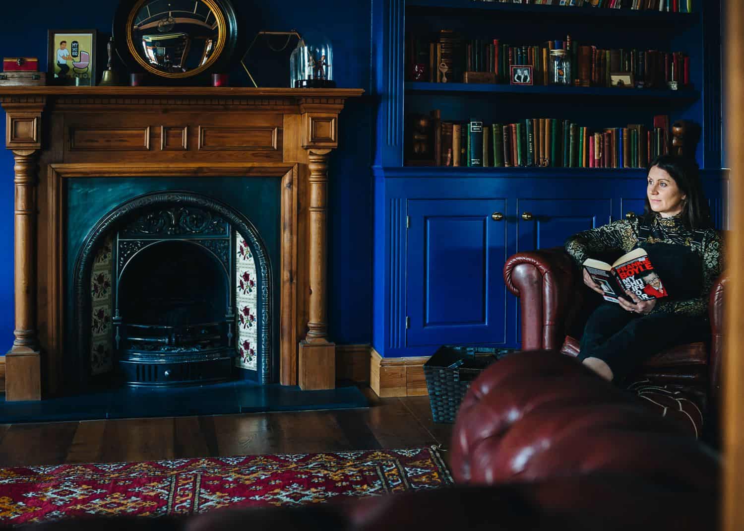 An author sits in a deep leather chair in her blue-painted library reading a book