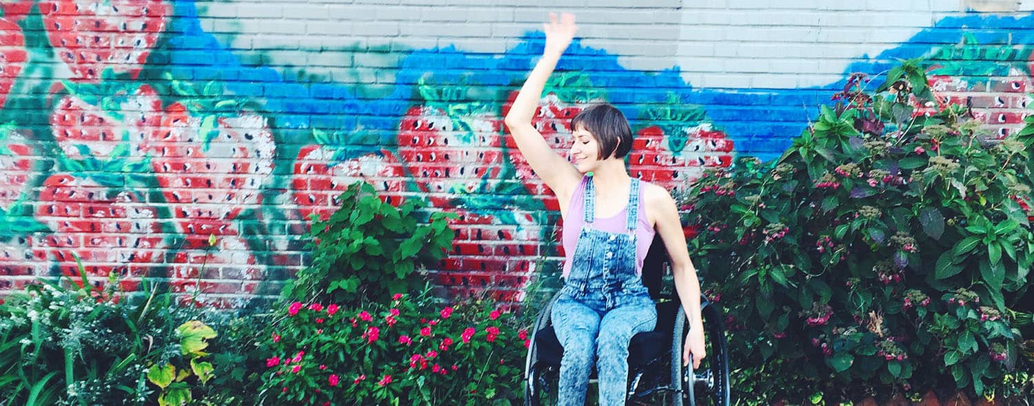 Writer Rebekah Taussig dances joyfully in front of a floral mural while sitting in her wheelchair