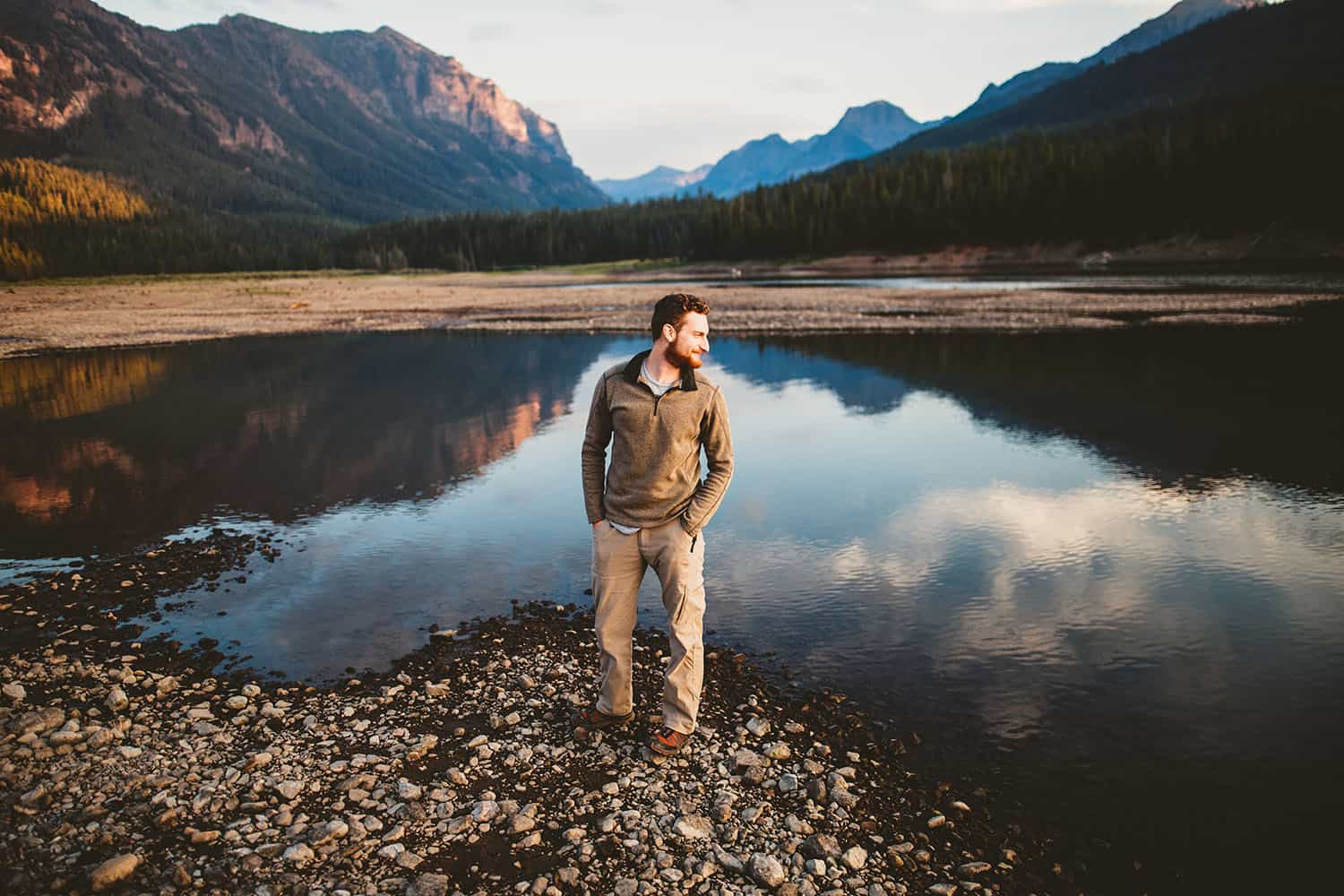 Color portrait of a caucasion man with a beard standing on the rocky shore of a lake with mountains in the background.