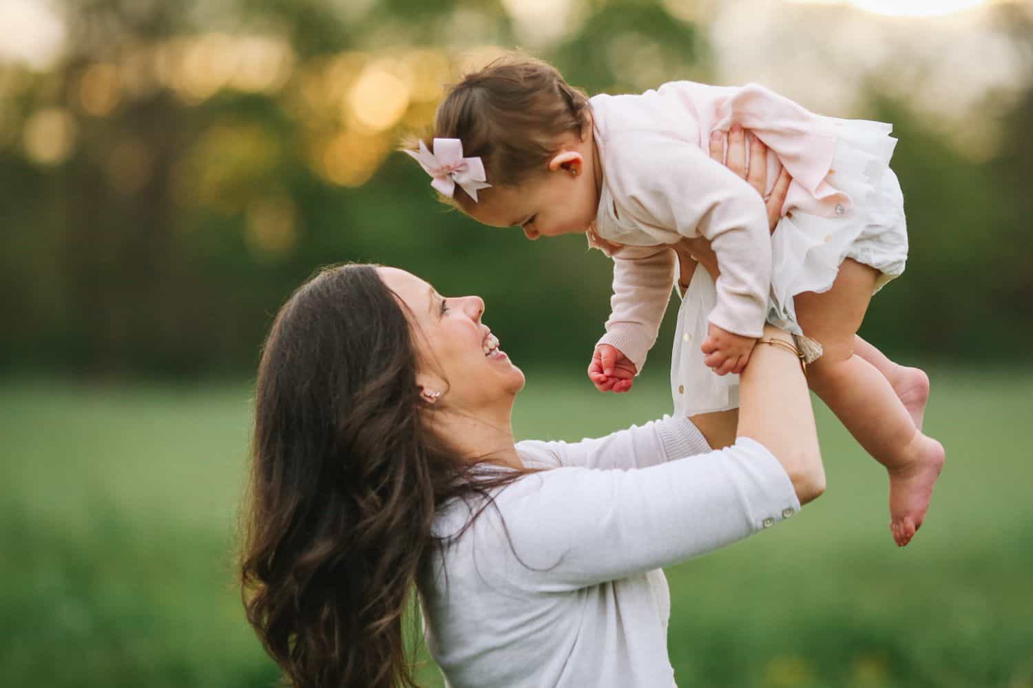 A mom in a field lifts her toddler daughter into the air at sunset