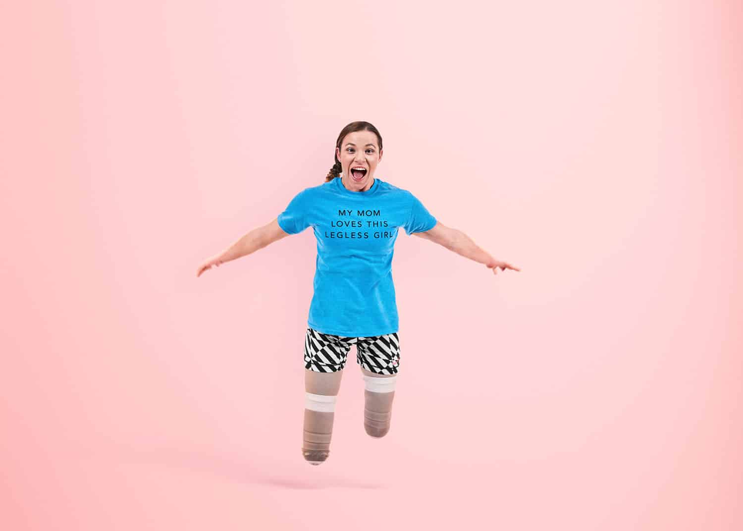 Paralympic gold medalist Oksana Masters leaps without her prosthetics on a pink background wearing a tee-shirt that says 