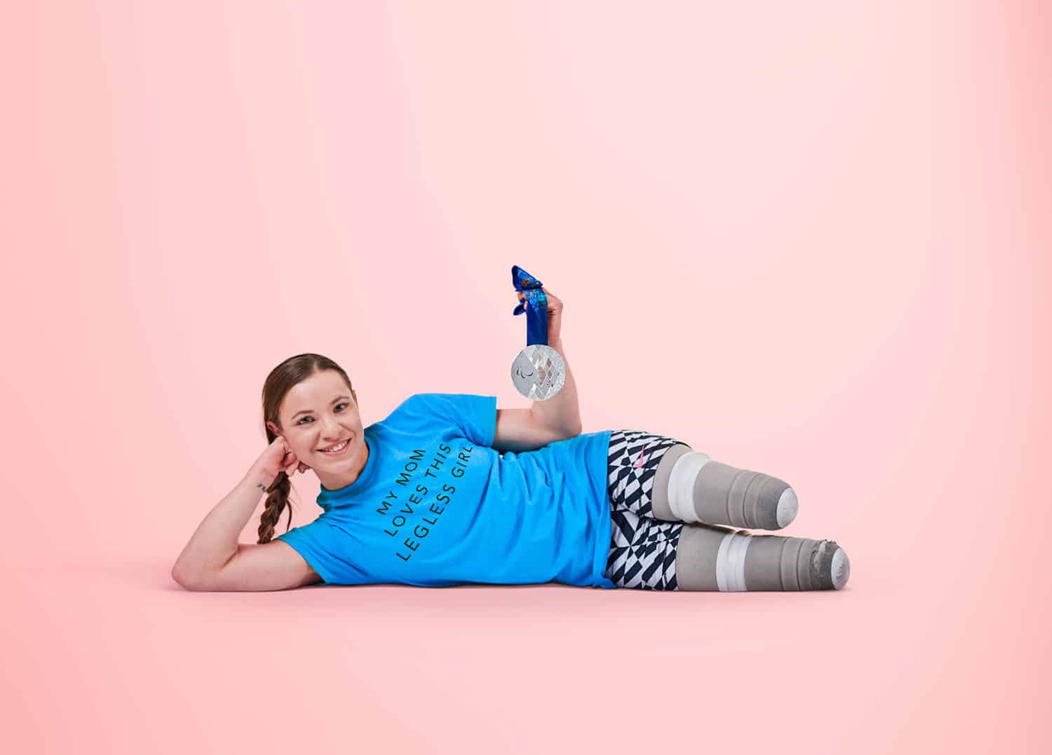 Paralympic gold medalist Oksana Masters reclines without her prosthetics on a pink background showing off her silver medal and wearing a tee-shirt that says 