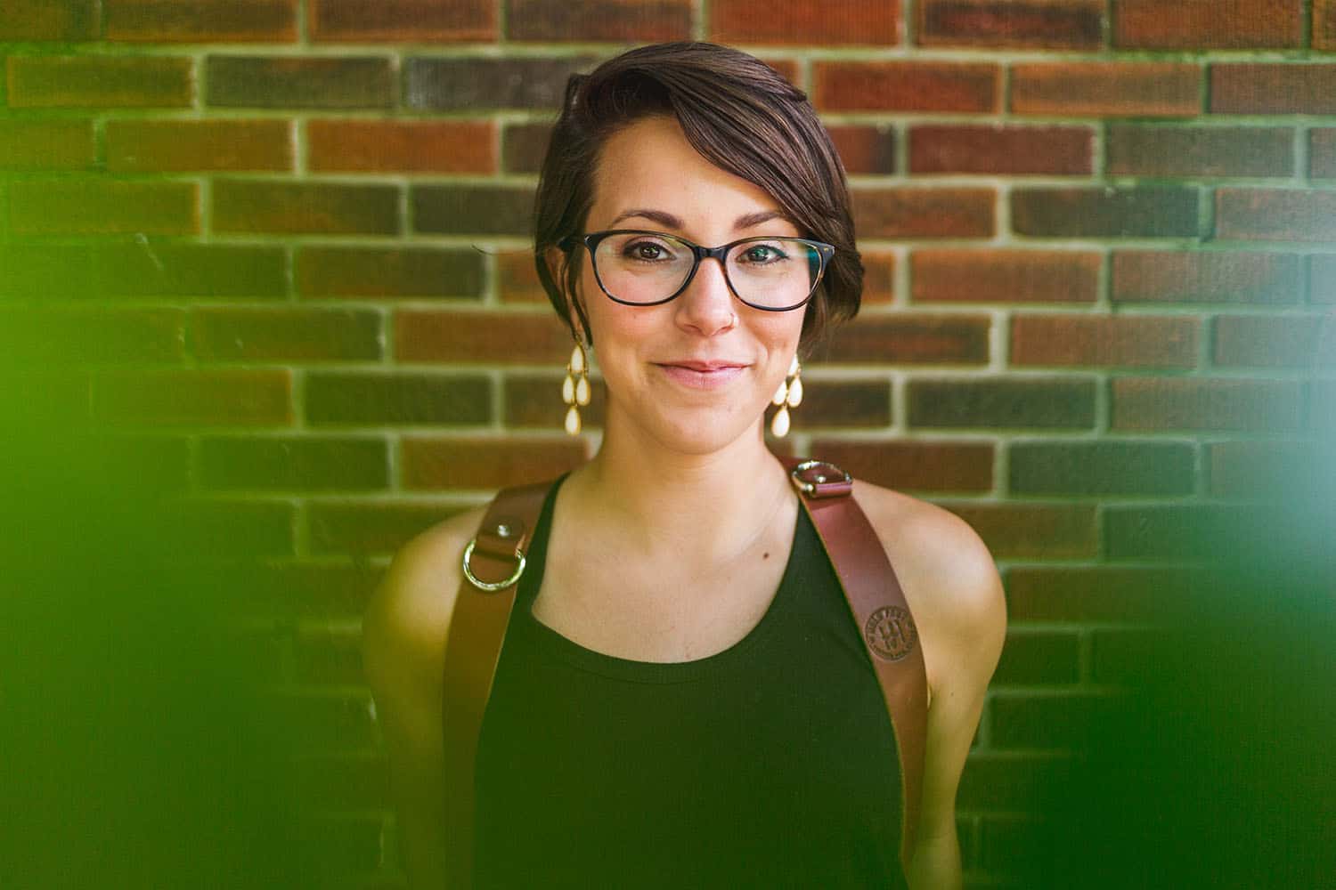 Photographer Mallory Regan, a caucasion woman with short brunette hair, glasses, and gold earrings, stands against a brick backdrop framed by leaves. She is smiling and wearing a HoldFast harness, made for carrying multiple cameras.