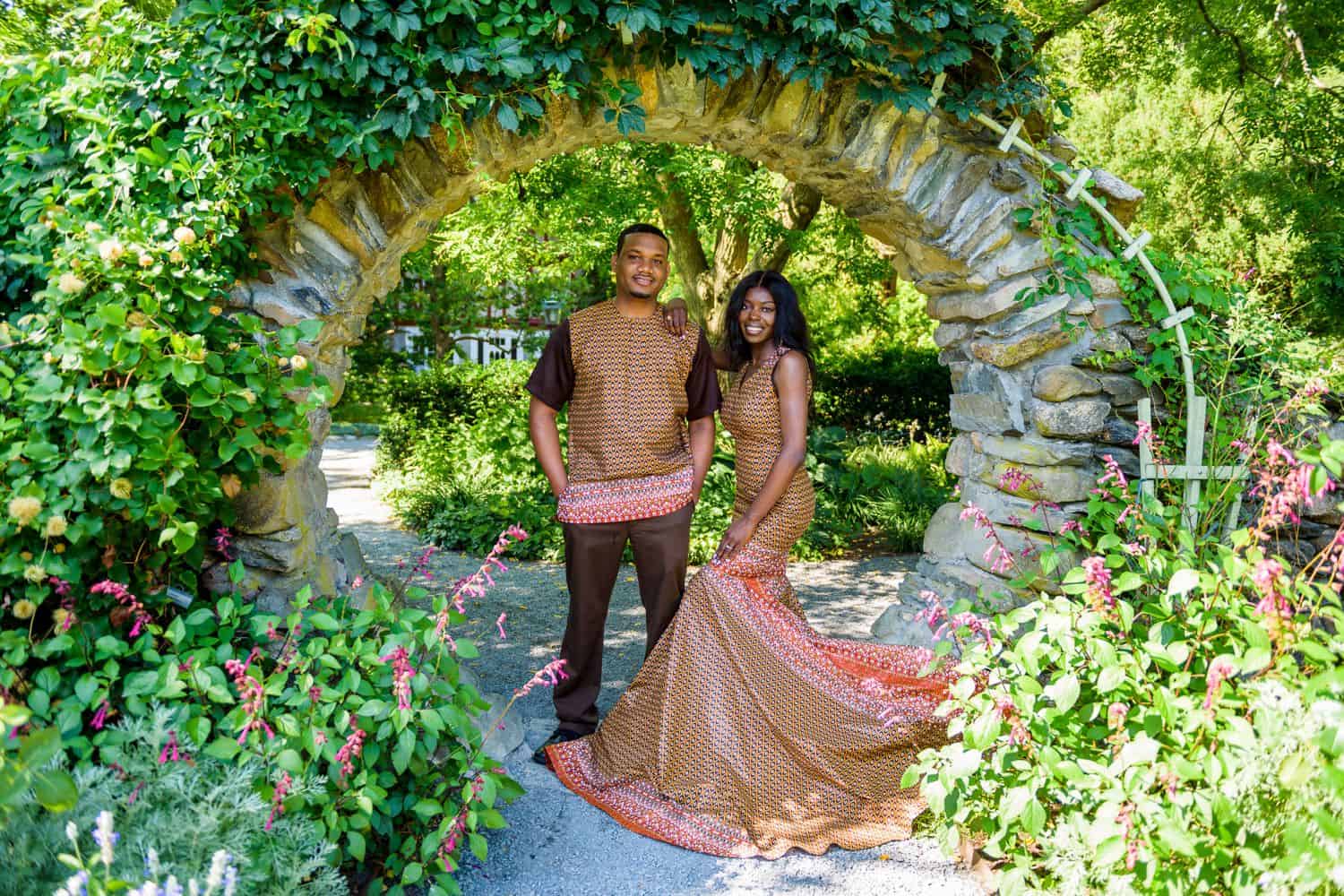 A Black couples poses in an ivy-covered archway wearing traditional African clothing