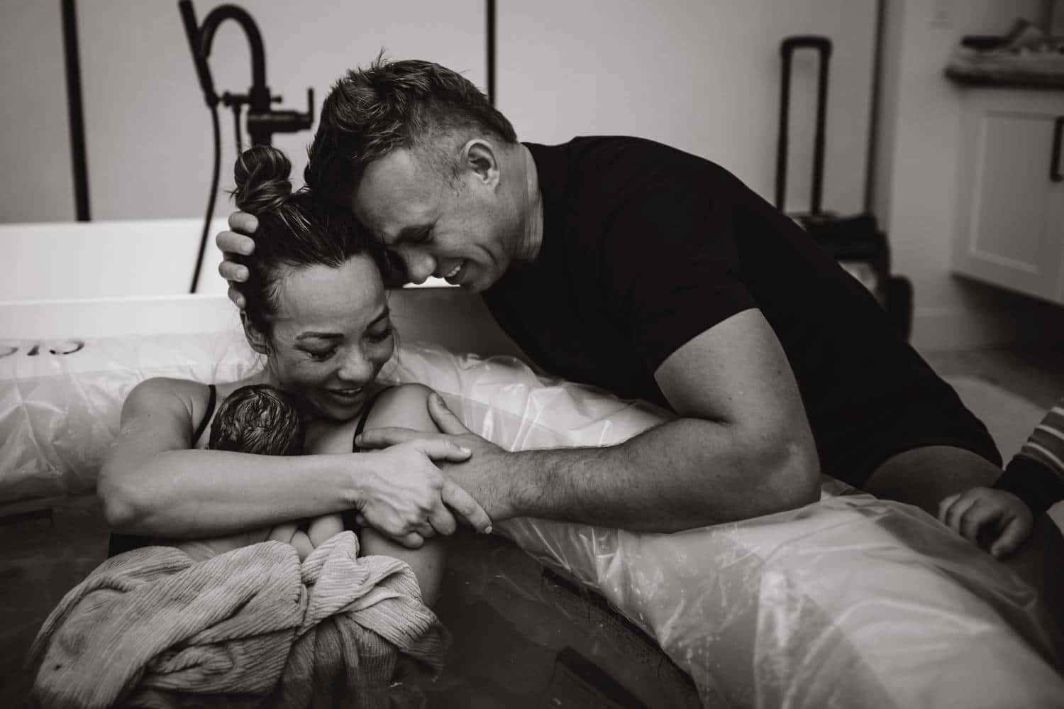 A partner embraces his post-natal wife and their newborn baby as the mother reclines in her home-birth tub.