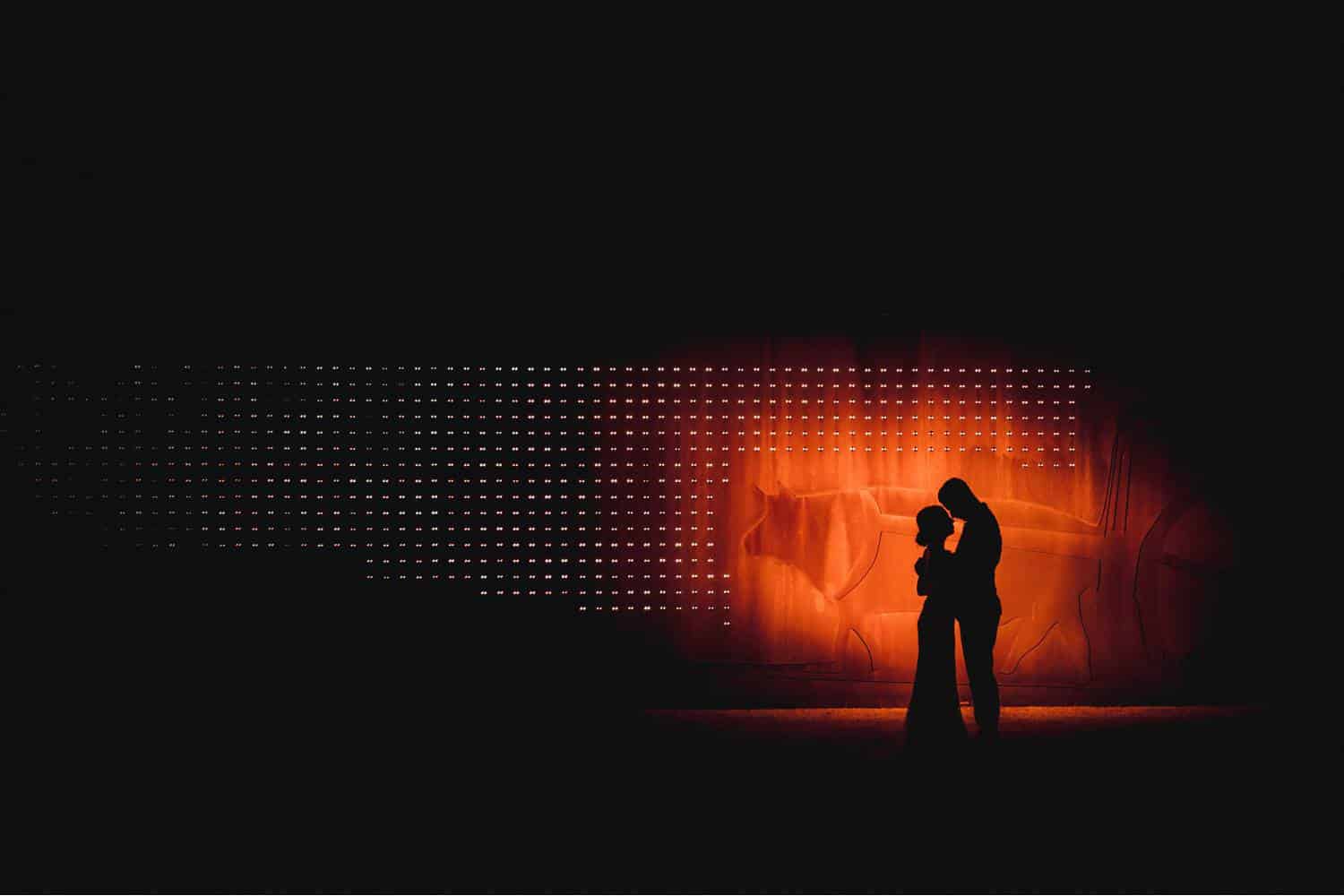 A couple is silhouetted against a glowing orange background. The photograph is framed so that the couple is highlighted on the right side of the image, with dark night all around their glowing forms. Black & Gold Photography educates photographers on how to make wedding portraits using off-camera flash.