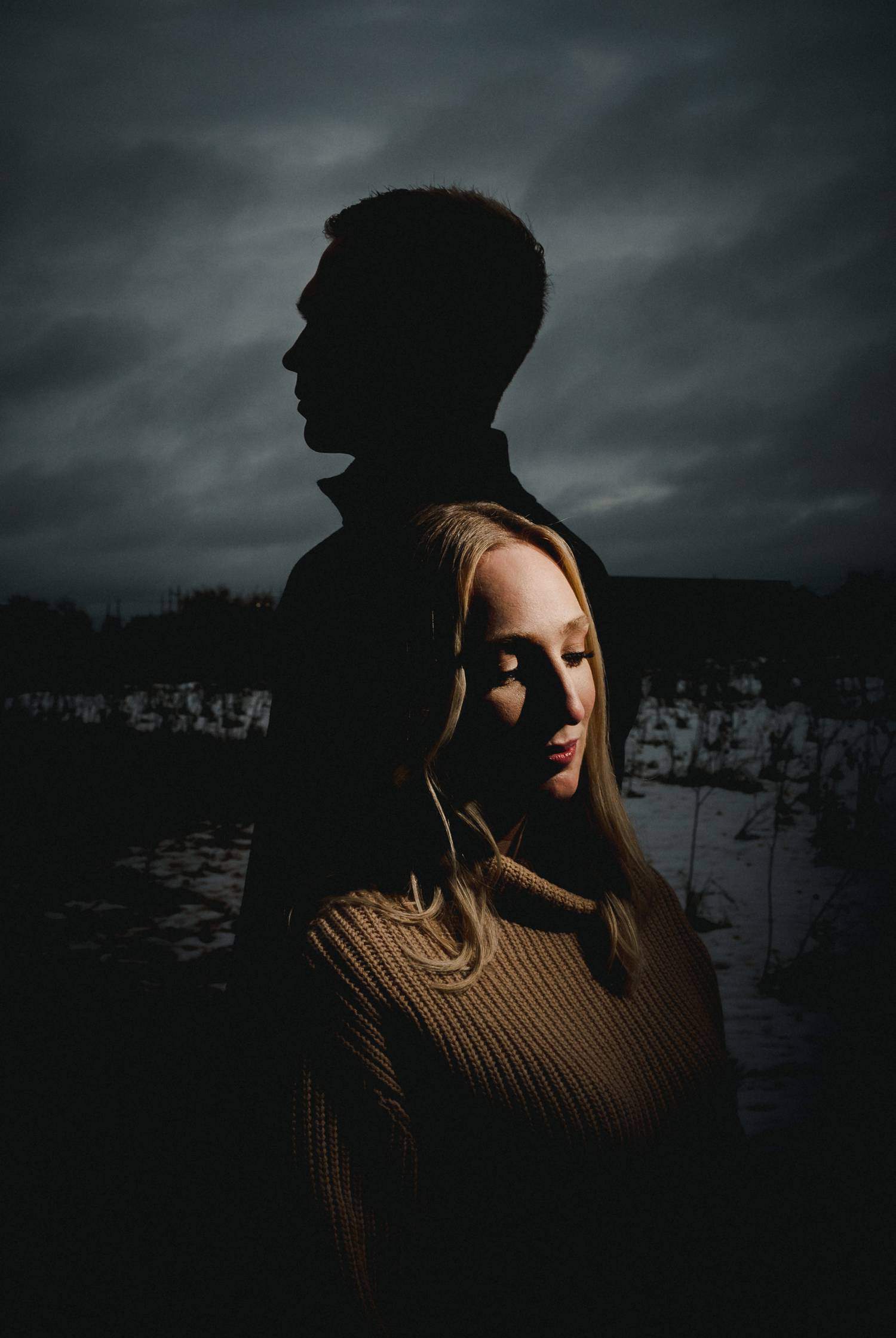 A man's silhouette appears against a cloudy winter sky. In front of his silhouette, a woman is light by a narrow beam of light. Black & Gold Photography uses unique lighting techniques to create wedding portraits using off-camera flash.