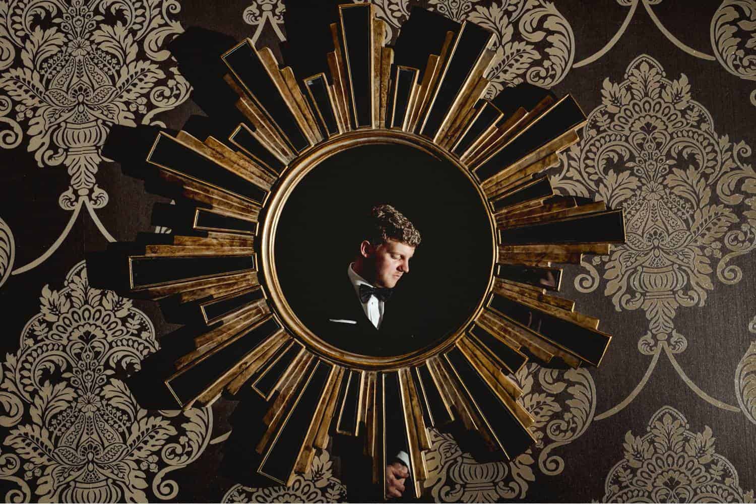 A modern starburst-shaped mirror hangs against old-fashioned wallpaper. Reflected in the mirror is a man in a tuxedo. His face is lit by a single flash. Black & Gold Photography creates these modern wedding portraits using off-camera flash.