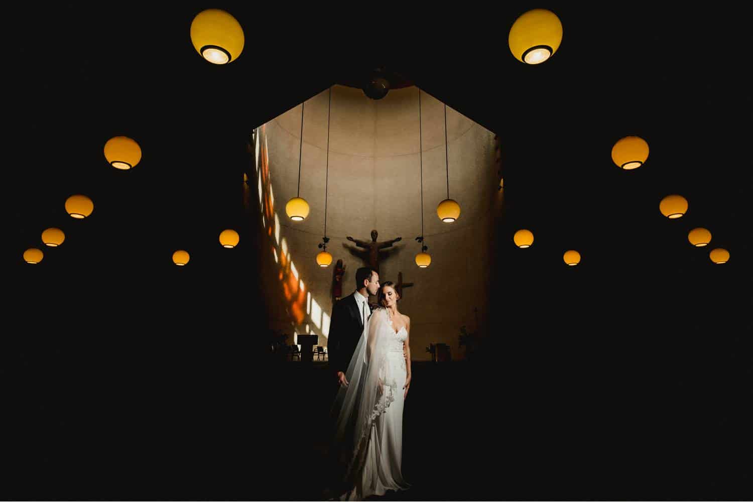 A bride wearing a long veil stands calmly beside a groom who looks solemnly at her face. Behind them, the sculpture of a saint is centered on a broad wall surrounded by round, hanging lamps. Black & Gold Photography makes stunning wedding portraits using off-camera flash.