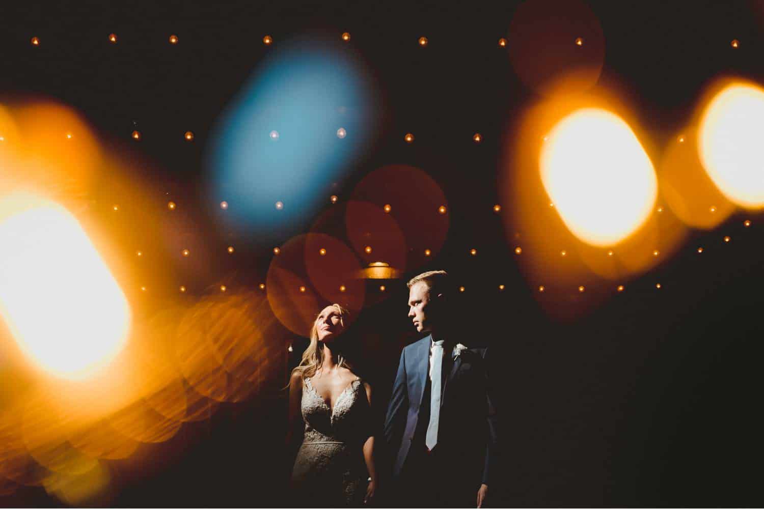 Black & Gold Photography created this double-exposure wedding portrait using off-camera flash. The bride stands at the center of the frame looking toward the bright light. The groom looks toward the light with his face angled toward the bride. They are surrounded by dream-like globes of light that were made using the double exposure technique.