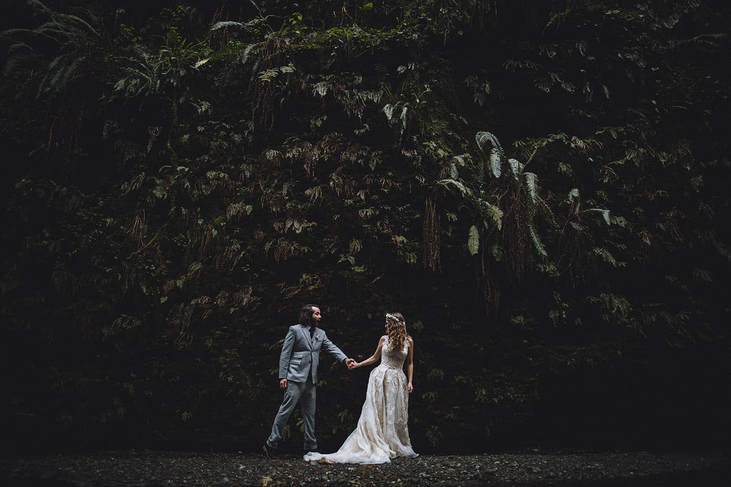 A bride leads her groom by the hand past a cliffside covered in ferns.