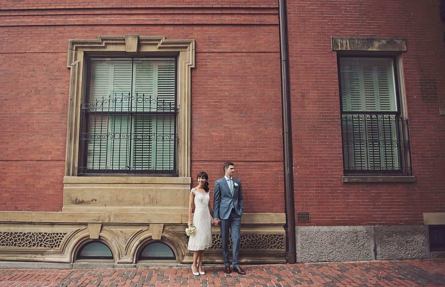 Bride and groom clasp hands in front of a tall brick wall. By Harris & Co.