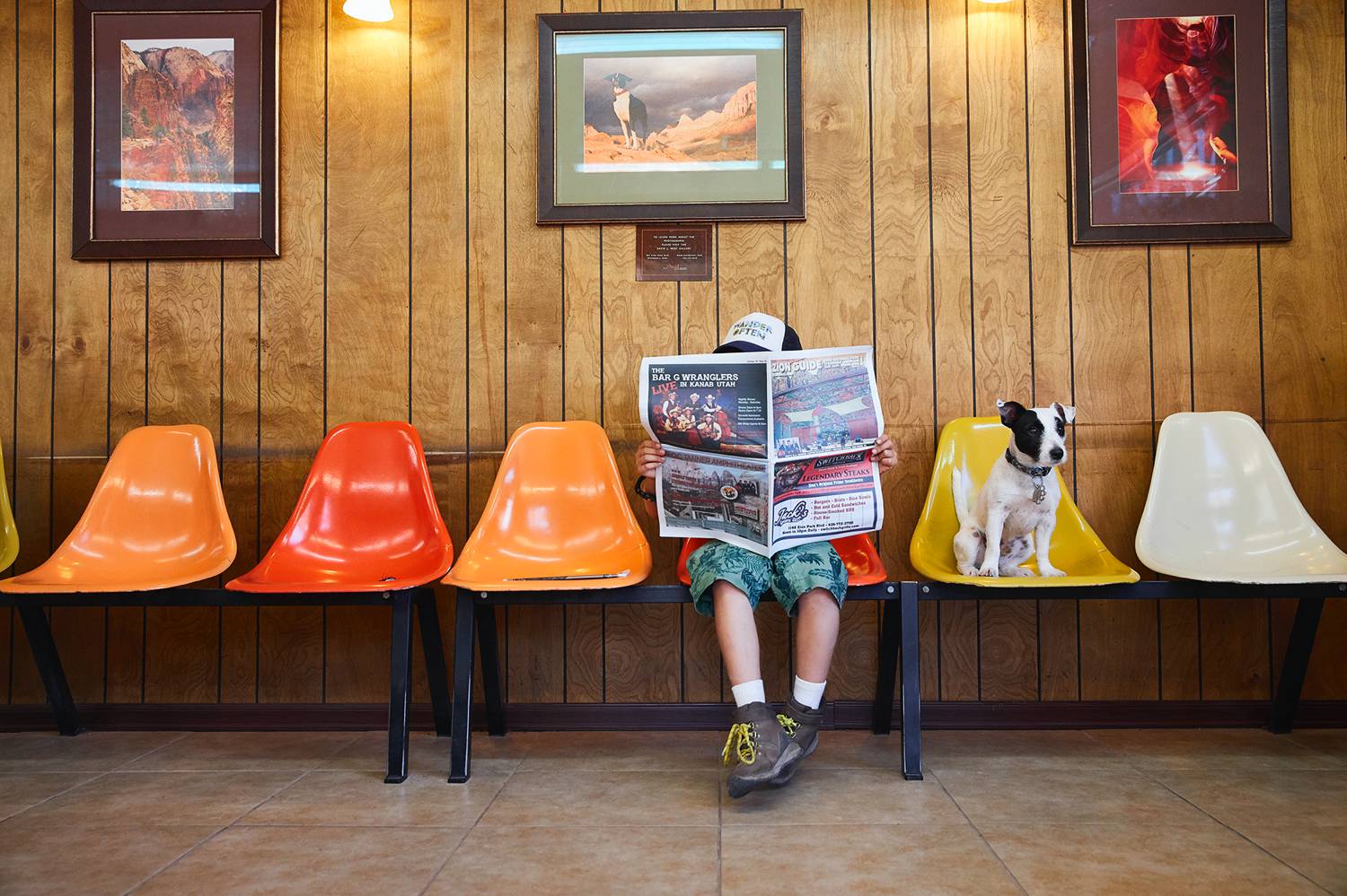 Photo: Candy Kennedy's documentary portrait shows a young boy sitting in a row of brightly colored chairs against a wood paneled wall. His face is shielded by the open newspaper he's holding. A small black and white dog sits in the chair next to him.