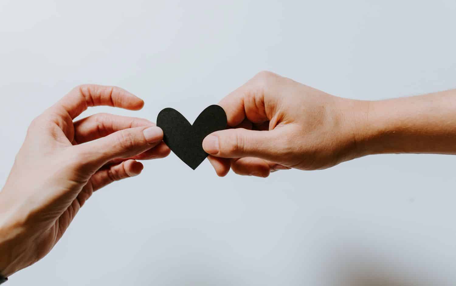 A person hands a paper heart to another person in front of a white backdrop.