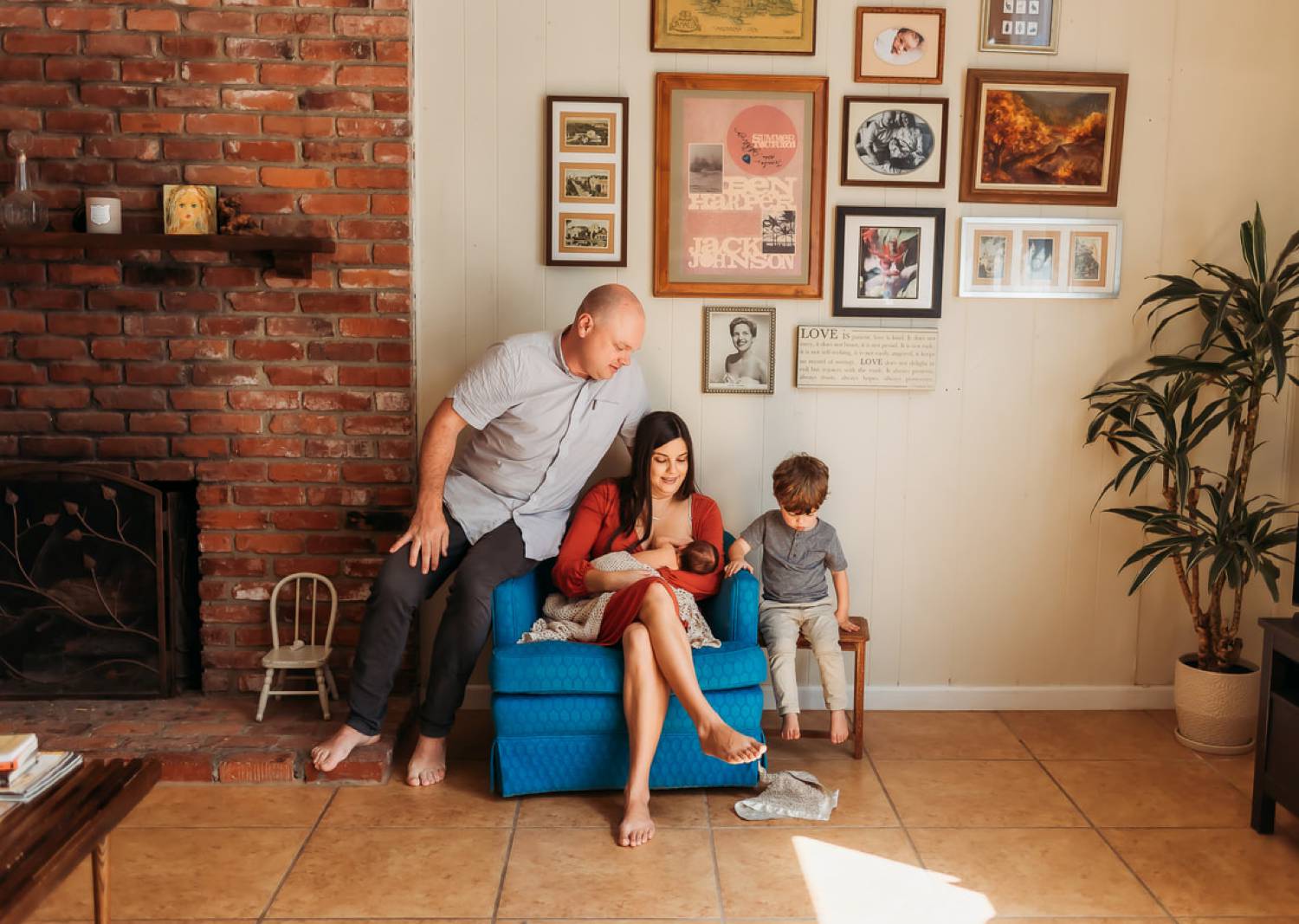 Photo: Charlotte Ani's in-home lifestyle newborn portrait depicts a mom sitting in a blue chair breastfeeding her infant. Her husband sits on the arm of the chair beside her, and their young son sits opposite his father on a small side table.