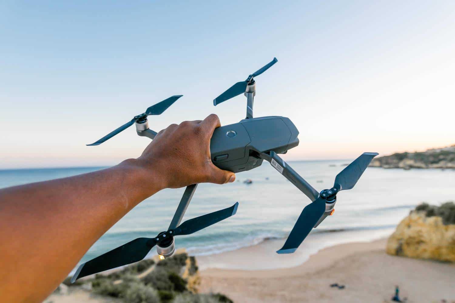 A hand is pictured holding a large drone camera in front of a beautiful ocean during a shoot by Humphrey Muleba