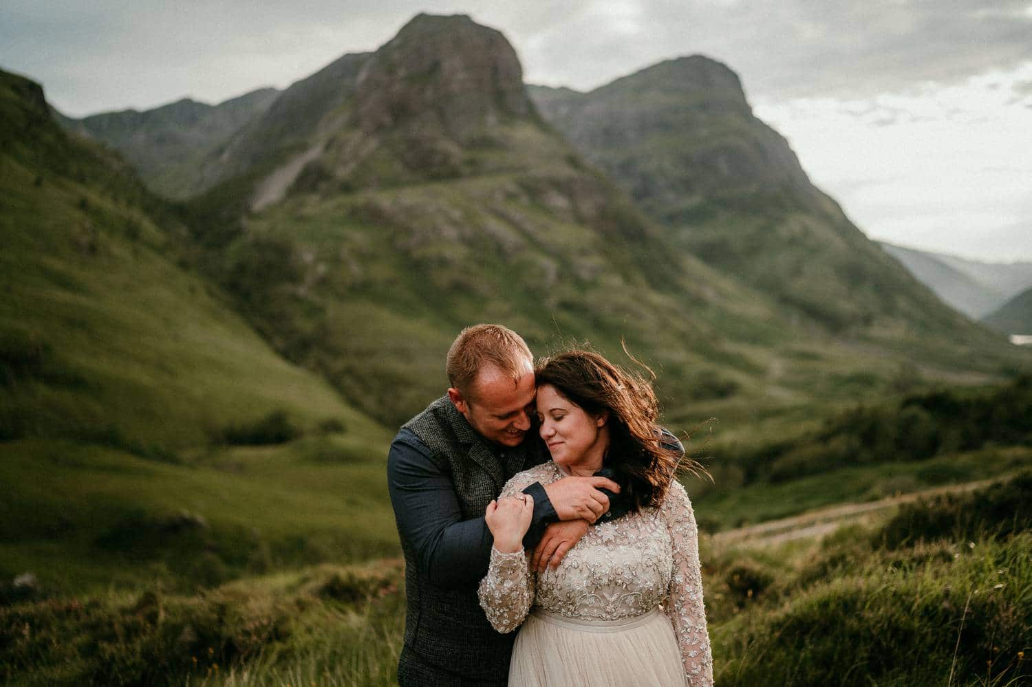 A groom stands behind his bride with his arms wrapped gently around her shoulders. She holds his arm and angles her face toward his in a loving embrace. Photographed by Rob Dight in the green hills of Ireland.