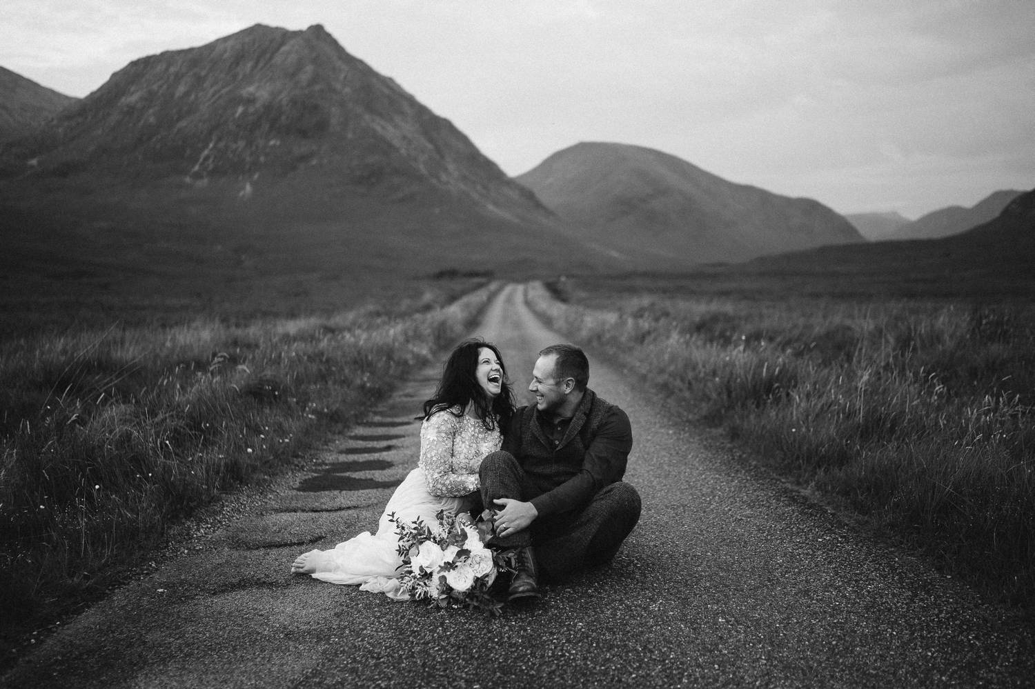 A bride and groom sit close together, laughing and embracing on an old Irish road. Black and white photograph by Rob Dight.