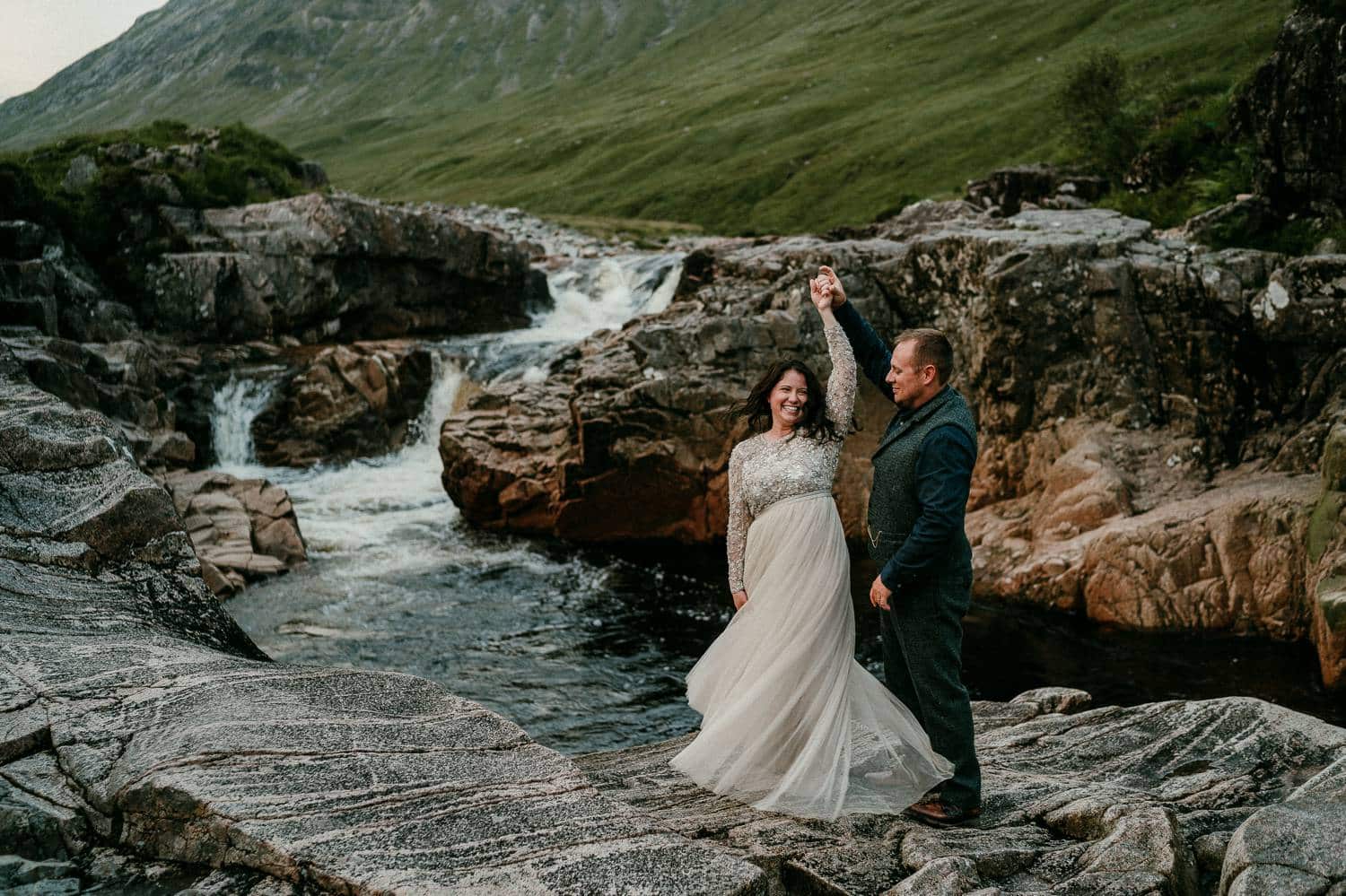 A groom spins his bride in a gentle dance move that sends her skirts twirling. They're standing beside the blue waters of an Irish creek. Photograph by Rob Dight.