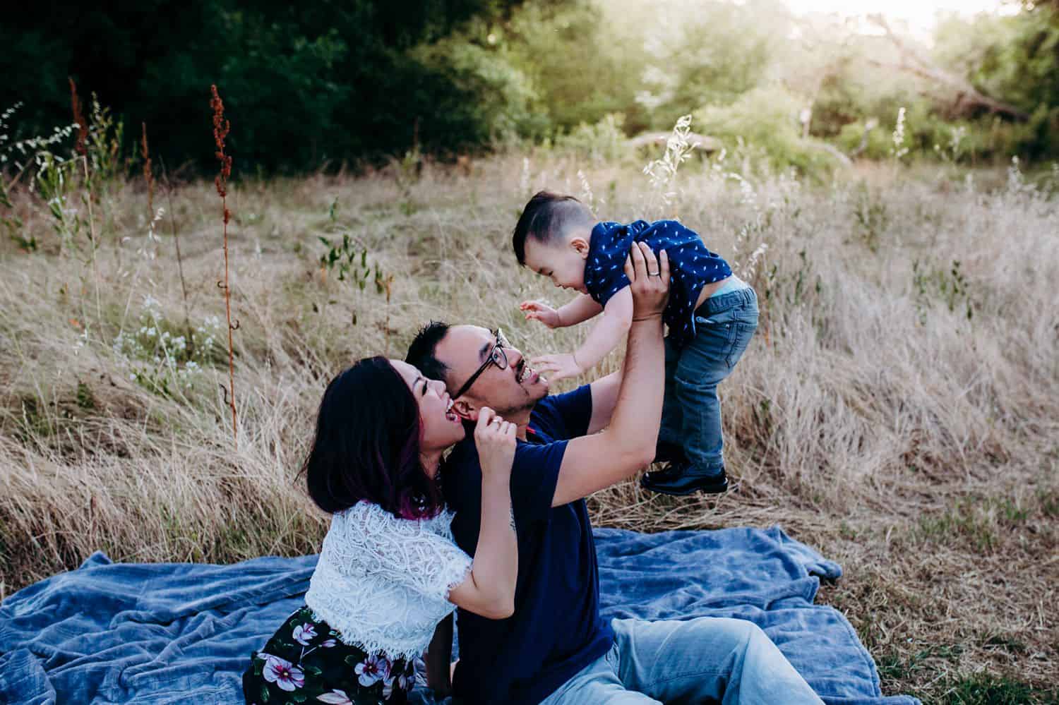 Two parents sit close together on a picnic blanket in the middle of a field while holding their toddler son up in the air and smiling.