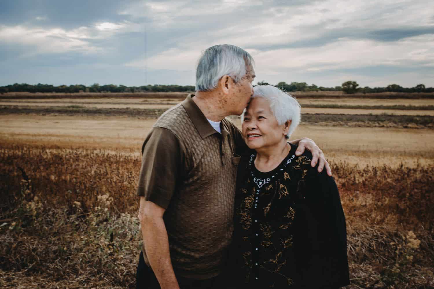 A grandfather kisses his wife on the forehead while standing in a field.