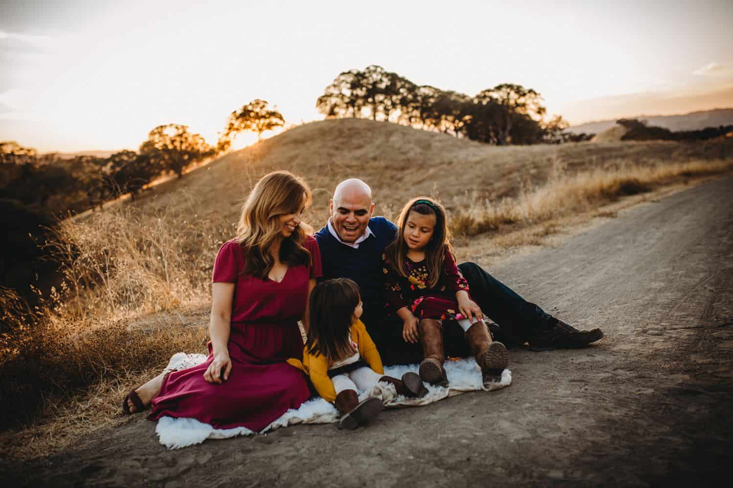 Two parents and their two young daughter sit on a picnic blanket in the middle of a field at sunset.