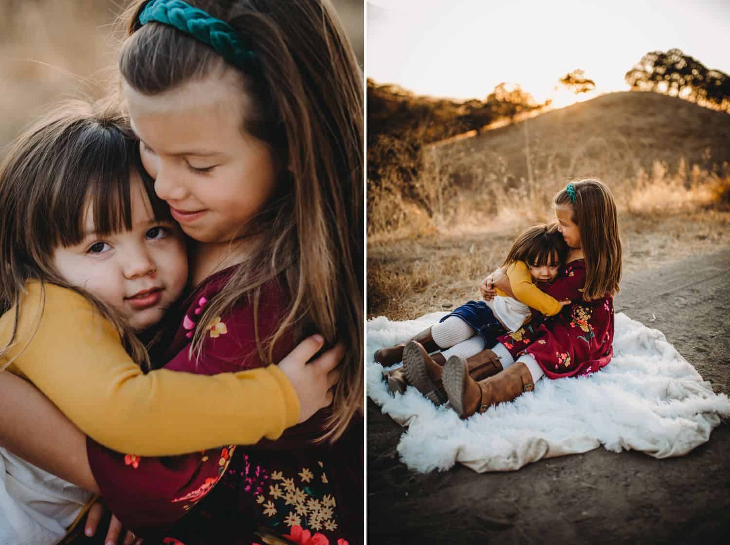 Two sisters hug while sitting on a furry blanket in the middle of a field at sunset.