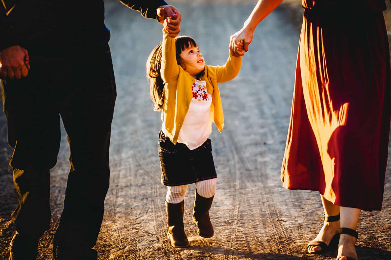 A little girl holds her parents hands and skips down a gravel road. The parents are only visible from the waist-down.