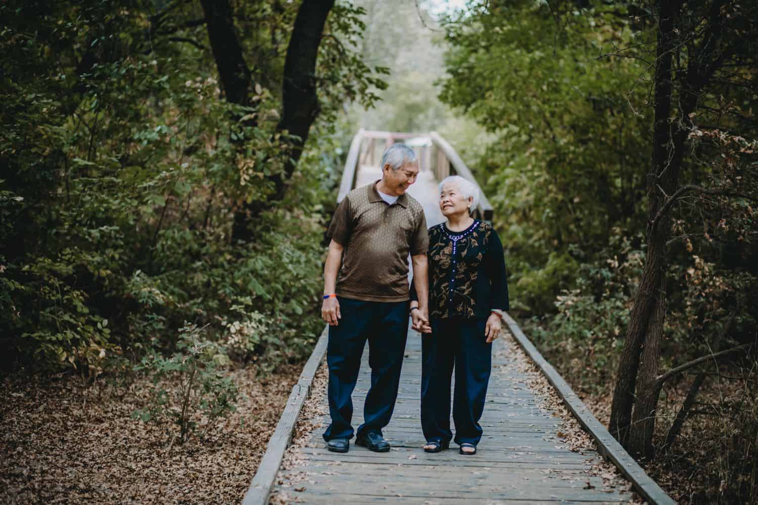 An elderly couple walks hand-in-hand down a forest boardwalk.