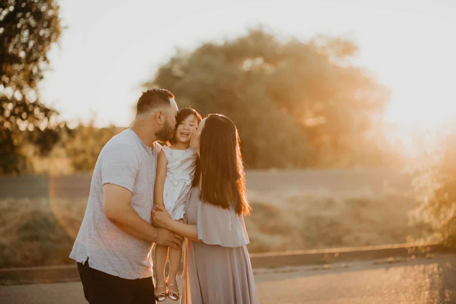 Two parents stand in a circle of sun flare. They're holding their young daughter between them and kissing her on both cheeks.