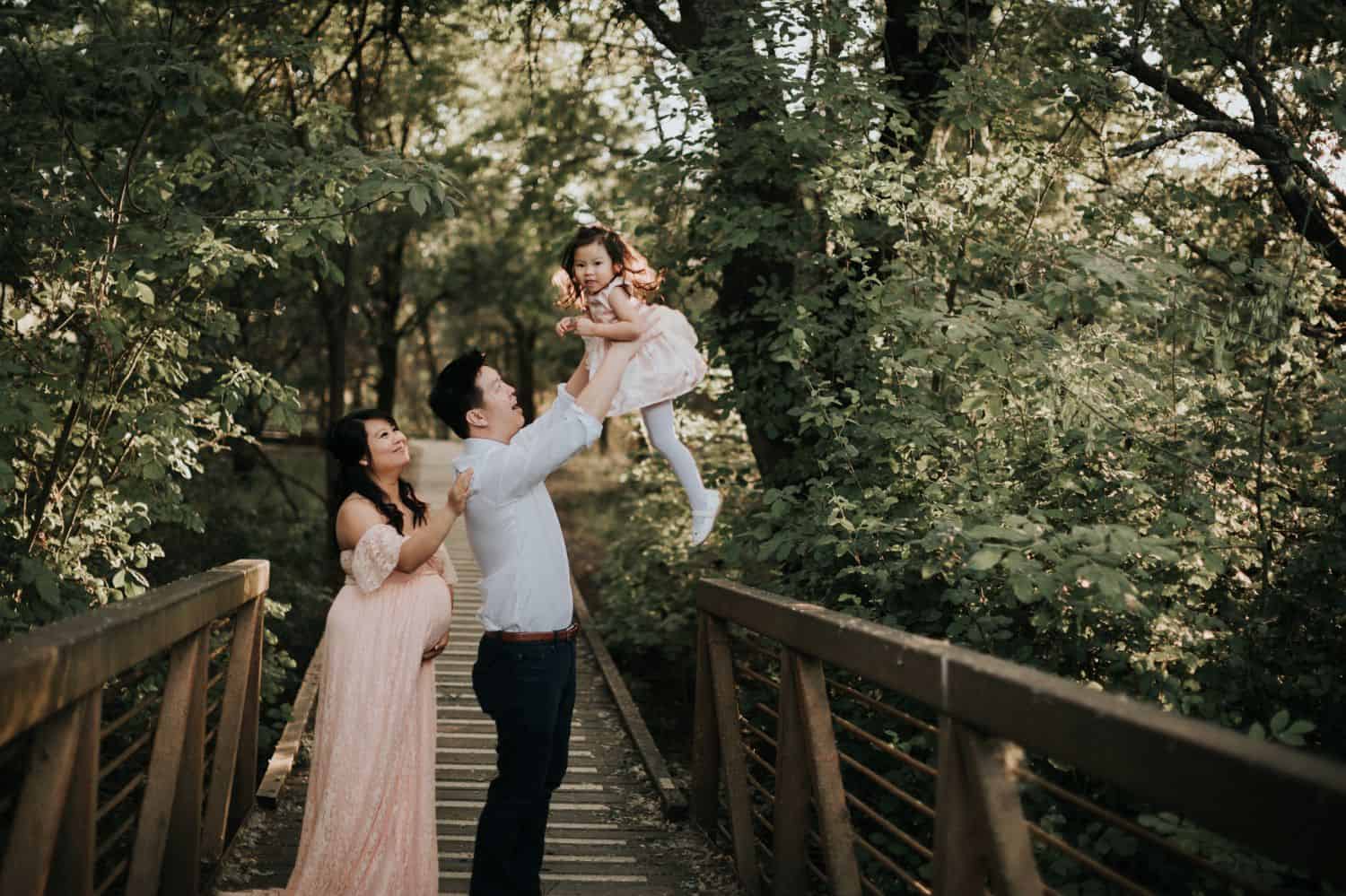 A pregnant mother stands behind her husband on a forest boardwalk. The father is holding his toddler daughter up in the air and laughing.