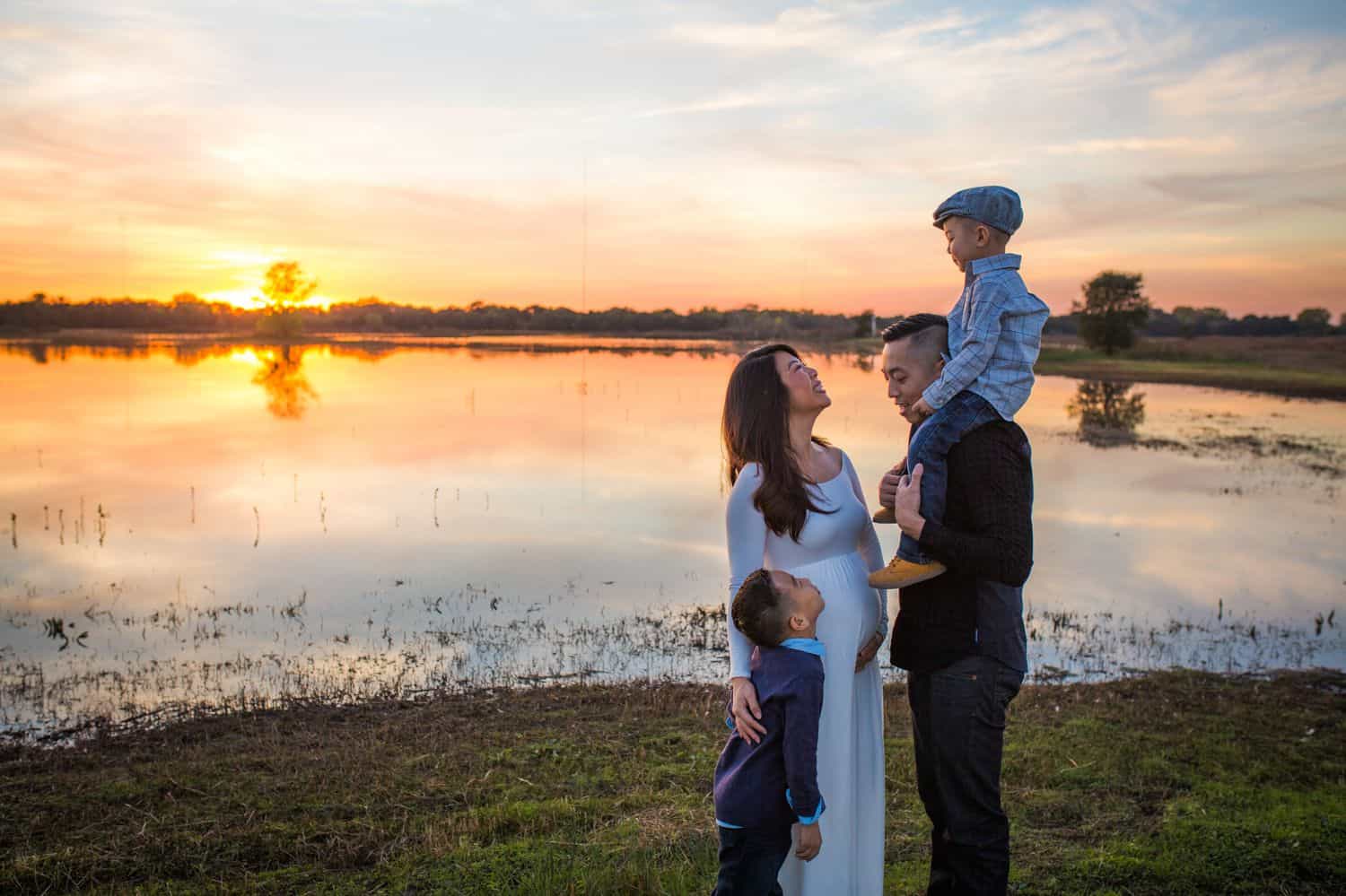 A pregnant mother and her husband stand with their two young sons beside a pond at sunset.