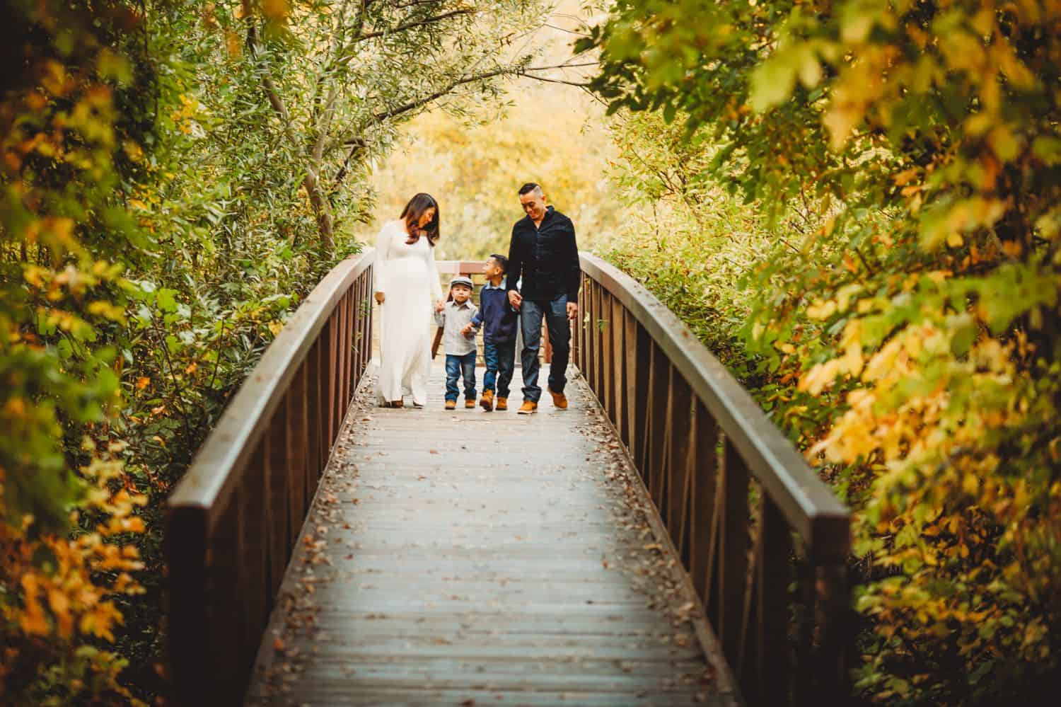 A pregnant mother in a flowing white dress walks down a forest boardwalk with her husband and two young sons. They are surrounded by fall colors.