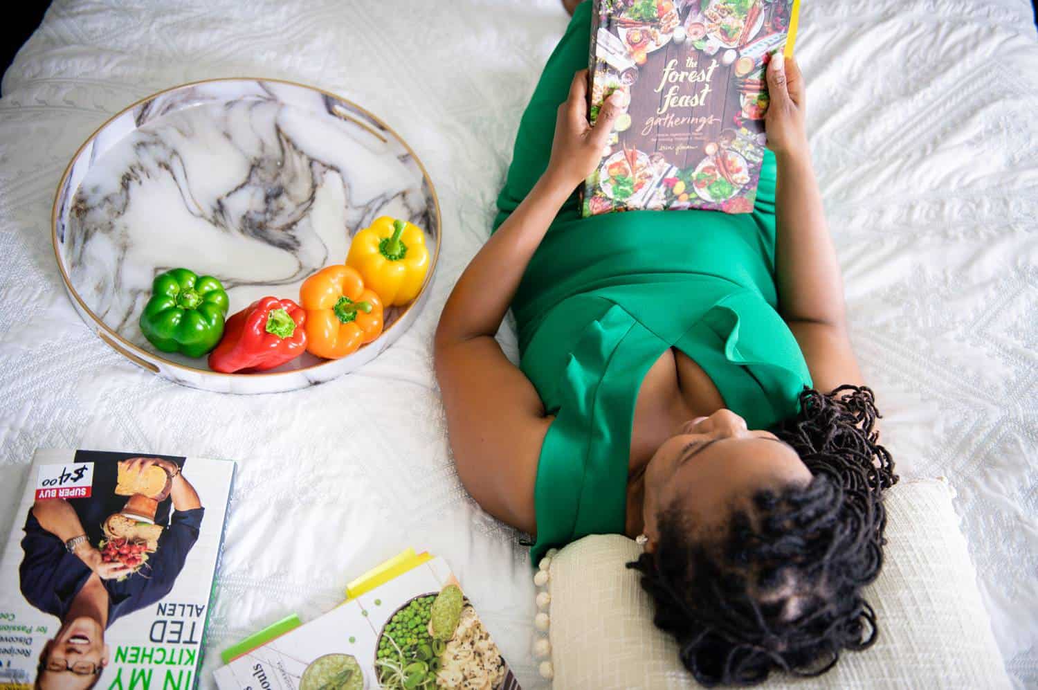 A Black woman in a satin, emerald green dress lies on a white bed reading a food magazine. Beside her is a marble tray of fruits and vegetables, along with a small stack of recipe magazines.