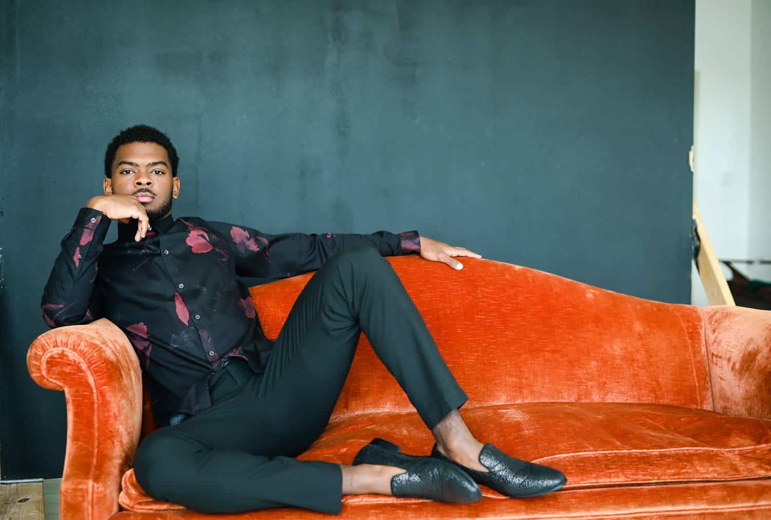 A dapper Black gentleman in black pants, a floral shirt, and alligator shoes sits casually on an orange velvet sofa with his feet propped up beside him. The photo is taken in a studio against a deep gray backdrop, and the man's rests his elbow on the sofa's arm so he can rest his chin in his hand.