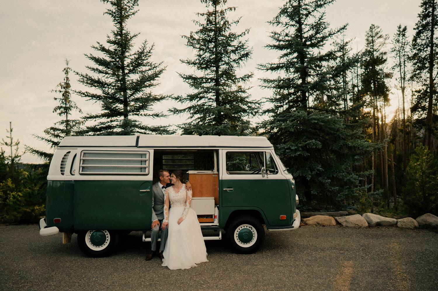 Customer Loyalty: By Nick Sparks, a camper van is parked in front of a stand of evergreens. A bride and groom sit in the open doorway.