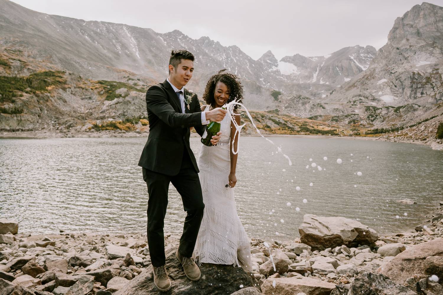 Customer Loyalty: By Nick Sparks, a bride and groom pop open a bottle of champagne beside a sparkling mountainside lake.