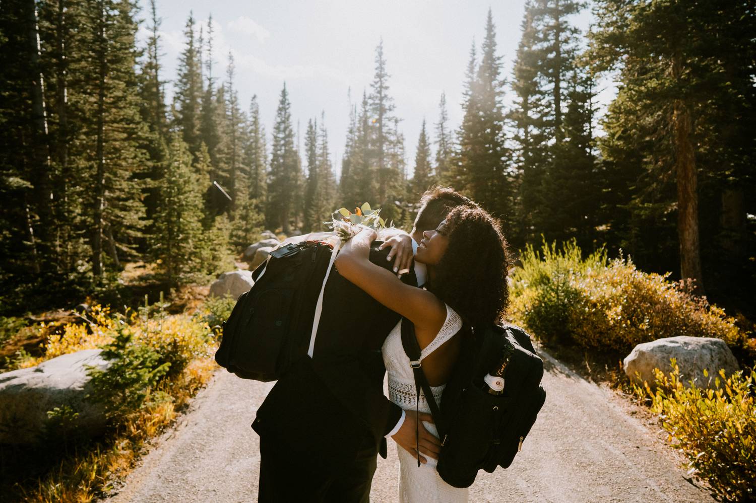 Customer Loyalty: By Nick Sparks, a bride and groom wearing backpacks hug in the middle of a mountain road with evergreens soaring behind them.