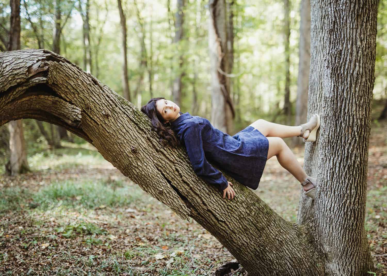 A high school senior girl lies in the fork of a tree wearing a denim dress and no shoes