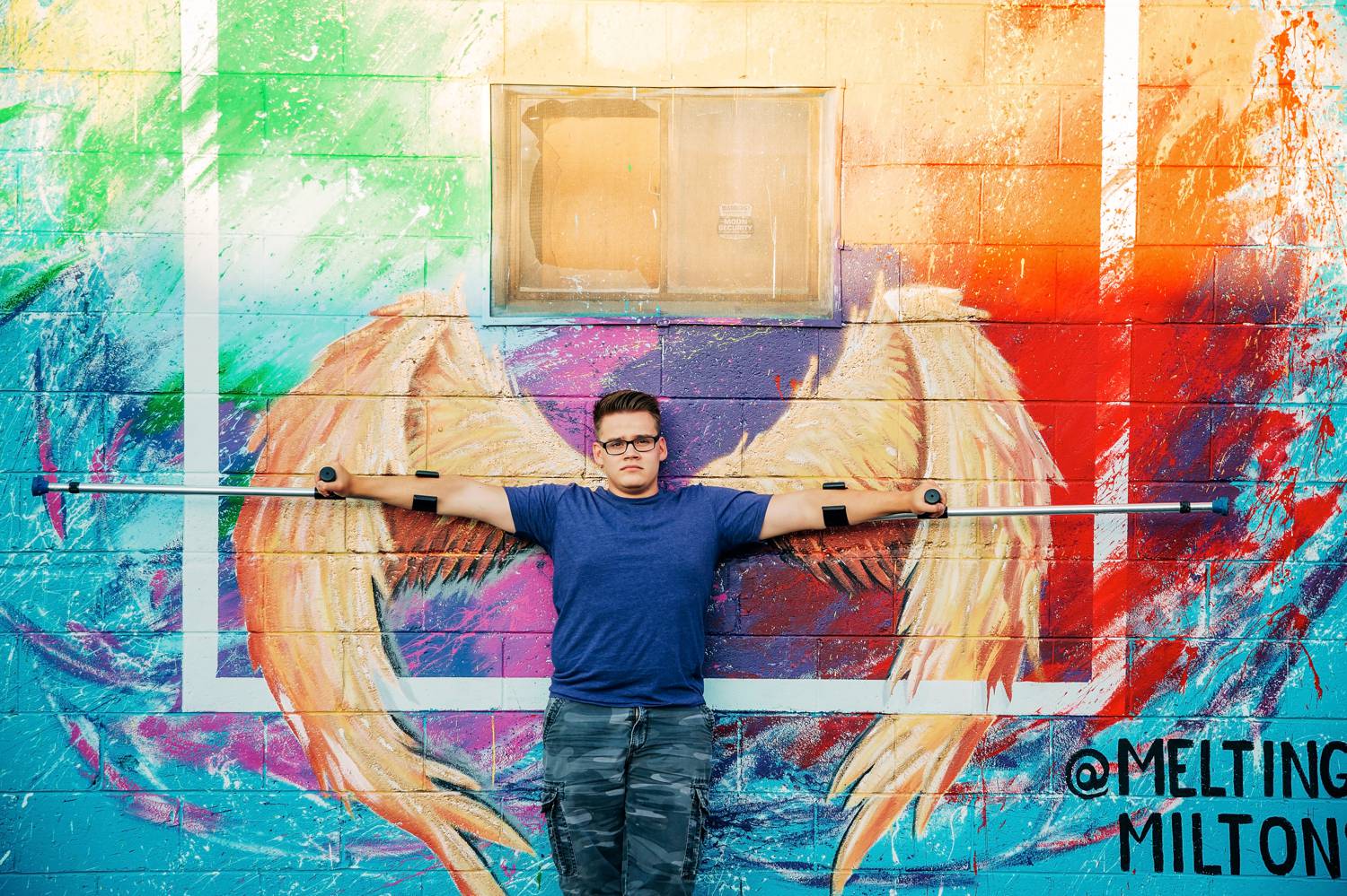 A young man in a blue shirt poses against a colorful mural of angel wings. He holds his arms outstretched with custom-fitted crutches extending from his hands, and the wings behind him make him appear angelic.